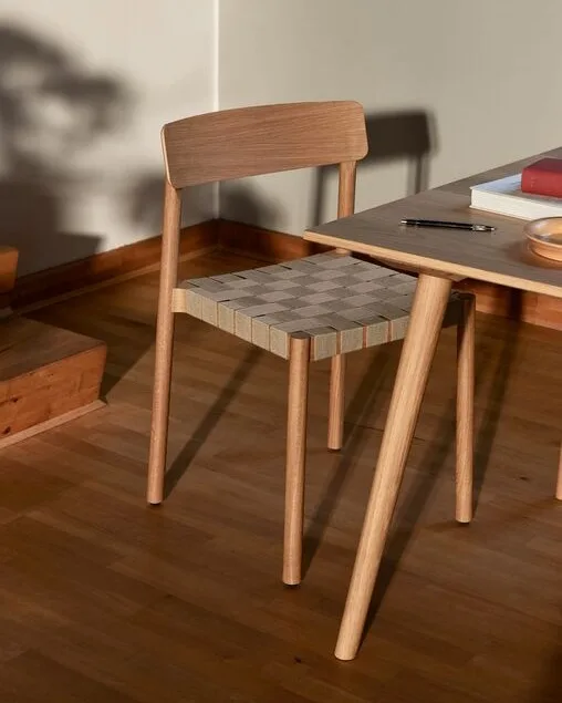 A light wood chair with a woven checkered seat next to a table with books and a pen.