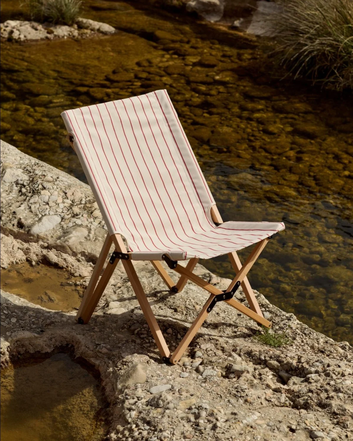 Foldable chair with a white and red striped fabric seat on a rocky bank next to a river.