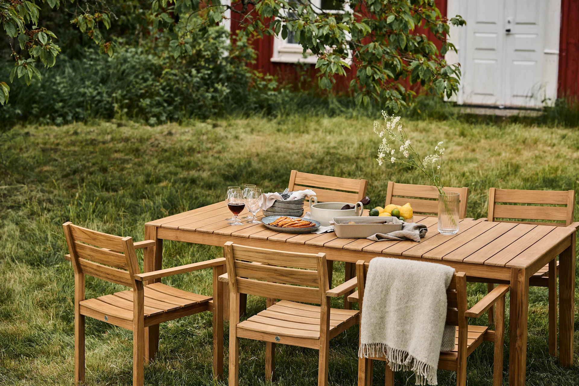 Outdoor dining set with wooden table and six chairs on a lawn, set with food, drinks, and a throw blanket, with a red house in the background.