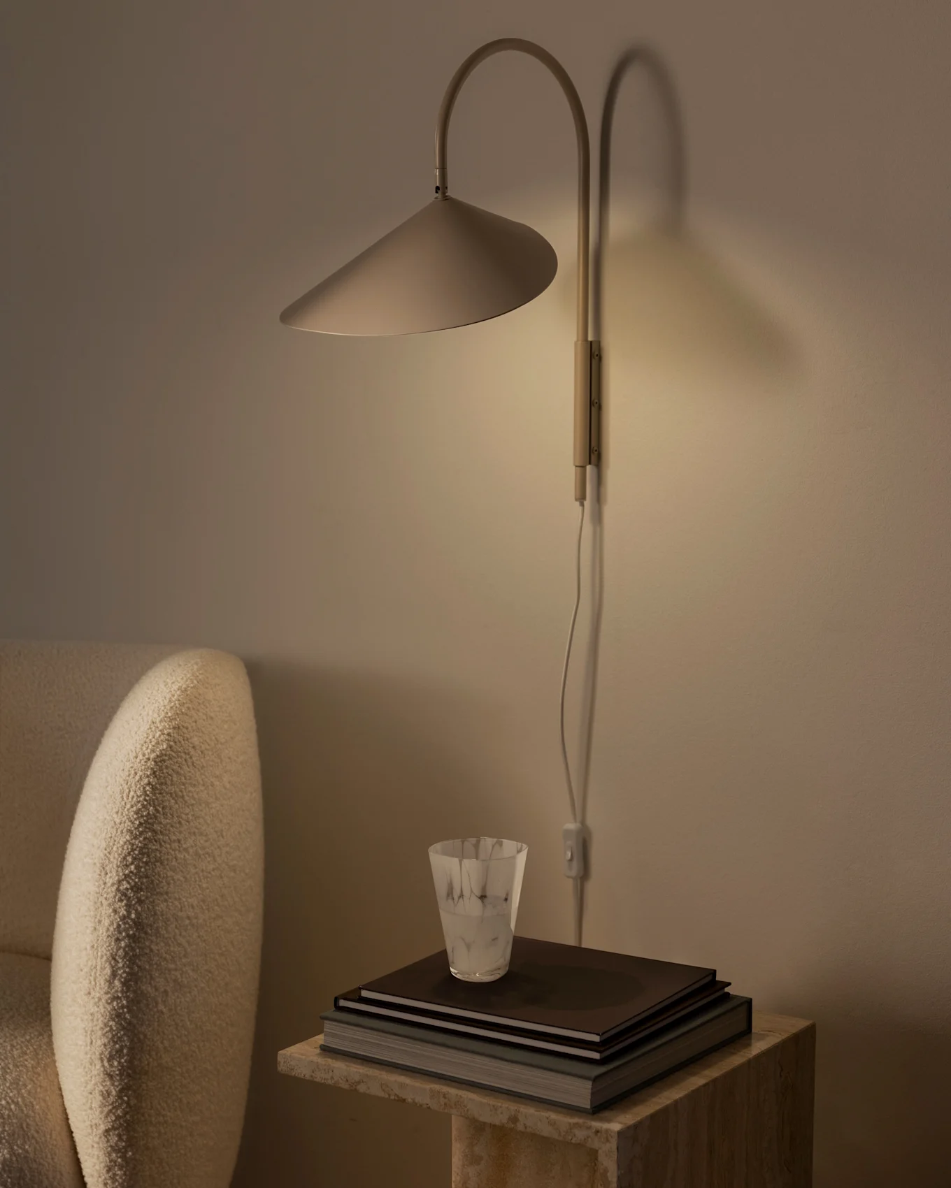 Warmly lit wall lamp with a conical shade above a stack of books and a textured glass on a stone side table, next to a beige armchair.