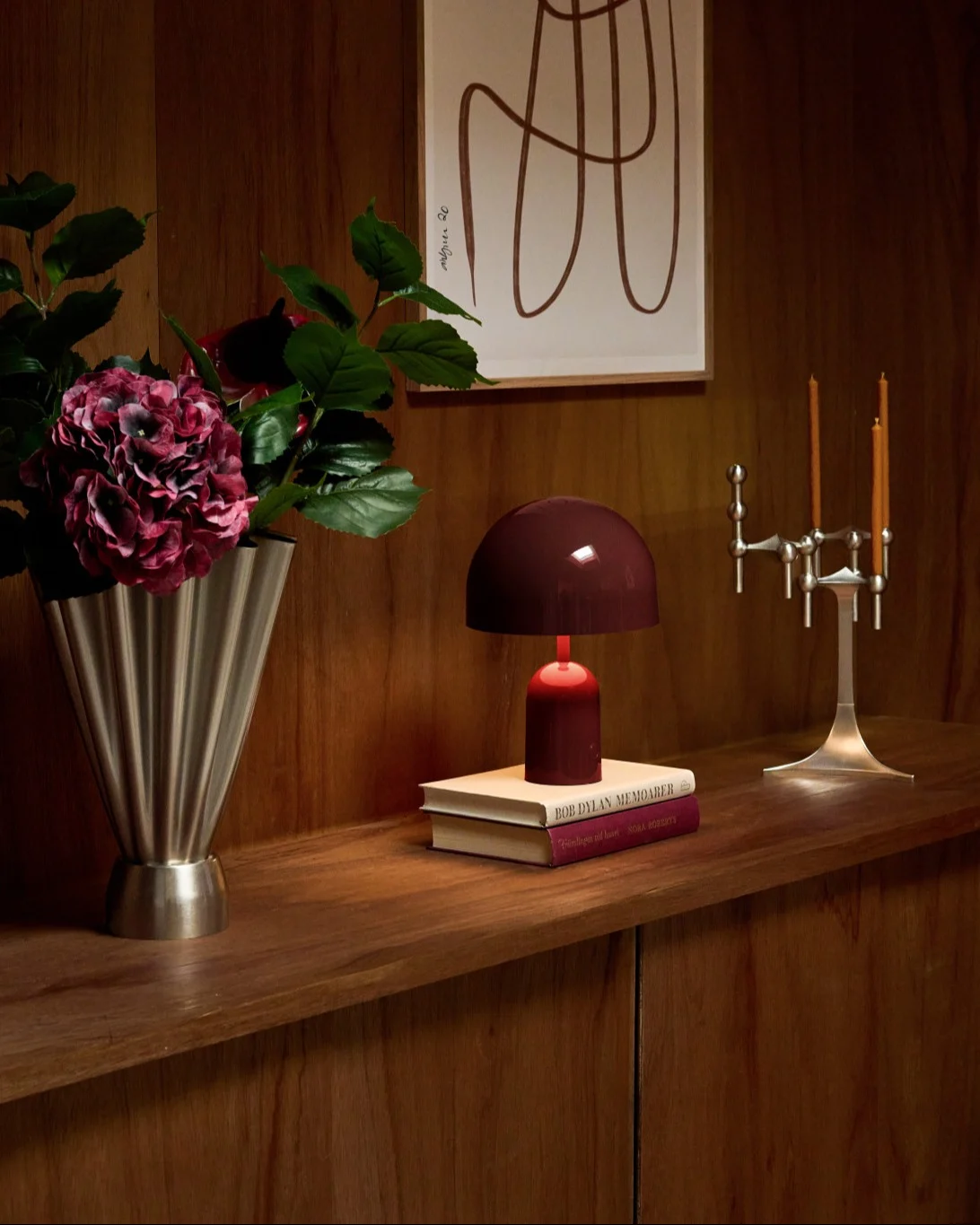 A cozy wooden shelf displays a silver fluted vase with deep red hydrangeas, a dark red lamp on books, and a silver candelabra.