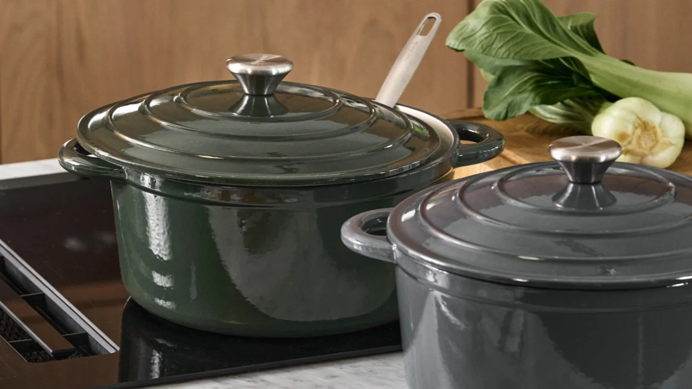 Two lidded cooking pots, one dark green and one dark gray, on an induction cooktop with bok choy in the background.