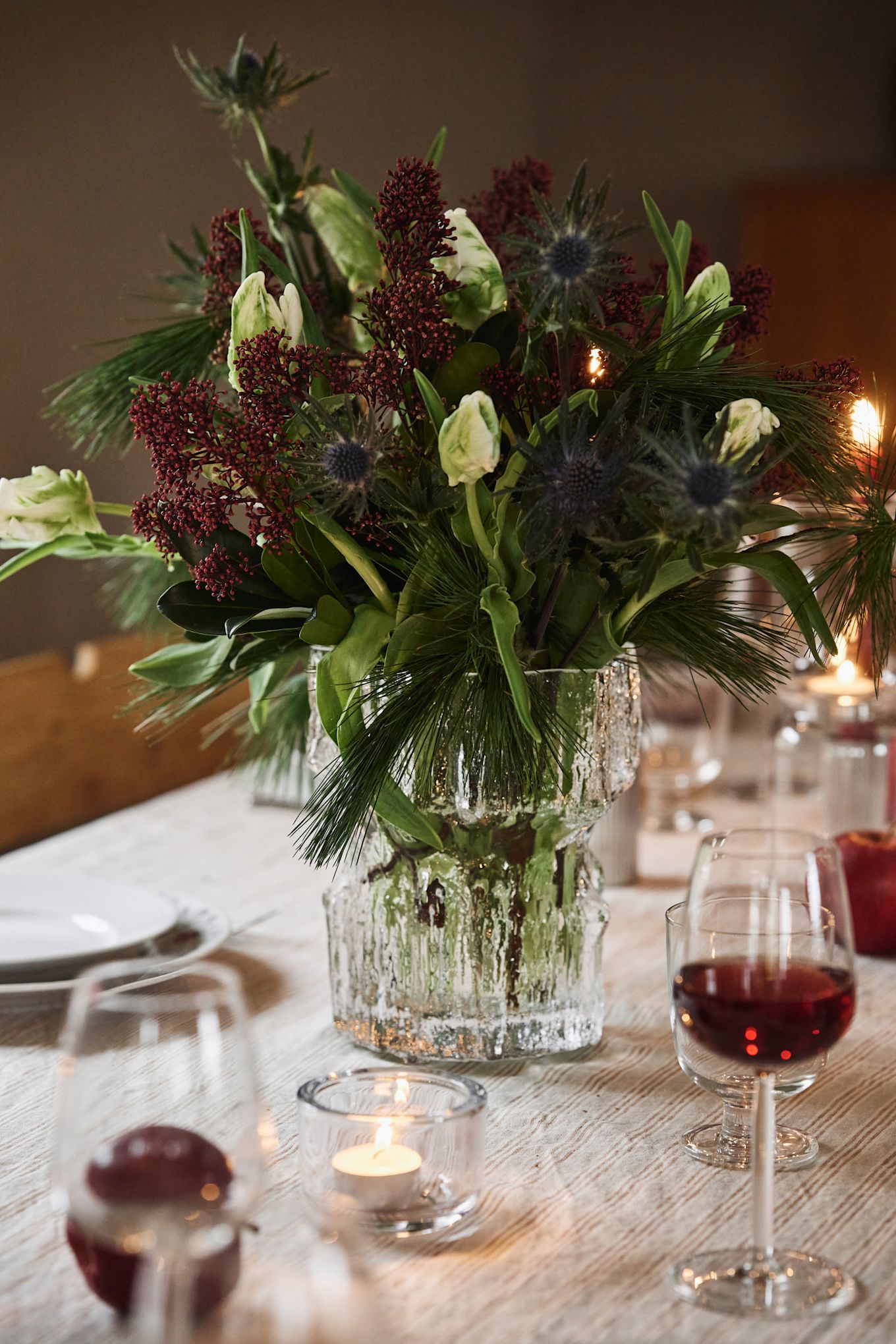 A textured glass vase with a wintery Christmas bouquet stands on a simple Christmas table setting. 