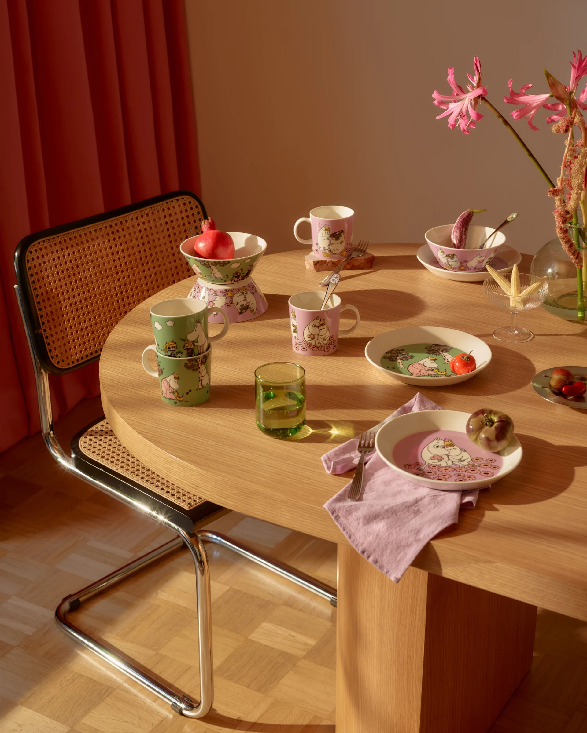 Dining table with pink and green Moomin-themed tableware, fresh fruits, a green glass, and pink flowers.