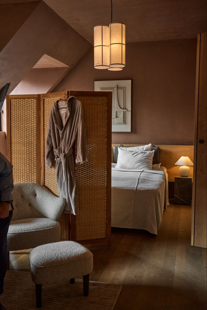 Design lovers should not miss an over night stay in Audo House. Here you see a cosy hotel room with a screen divider and a robe hanging in front of a bed. 