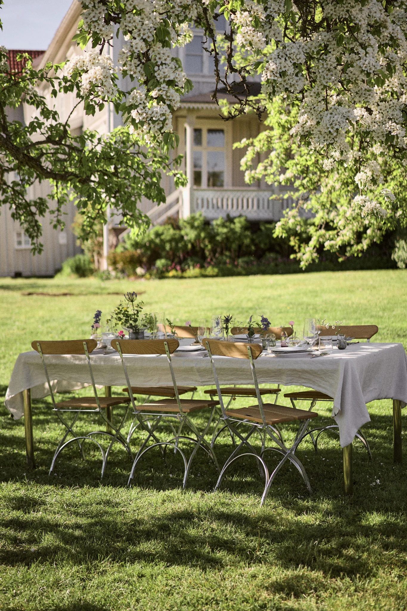 An outdoor dining table set with white tablecloth, plates, glasses, and floral decorations under a blossoming tree in a sunny garden.