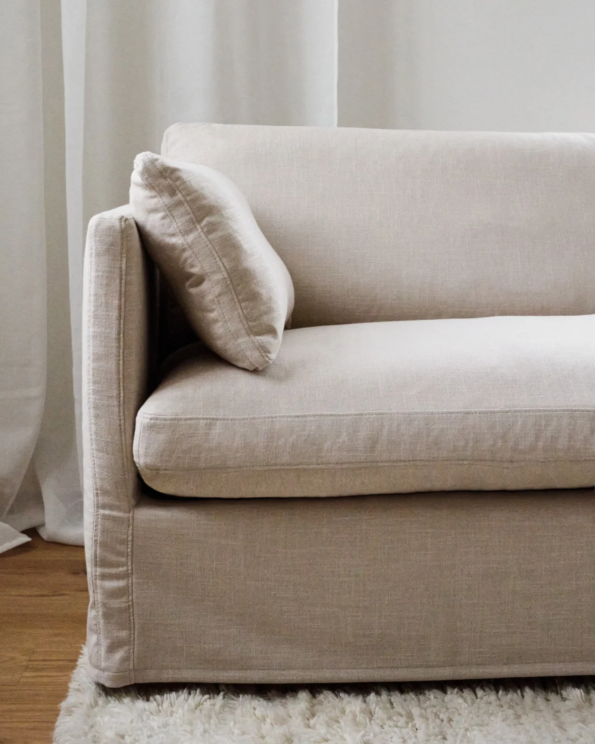 A close-up of a neutral-toned linen sofa with an armrest and a small pillow, sitting on a shaggy rug.