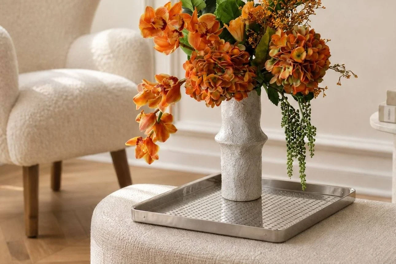 Vibrant orange flowers in a textured white vase on a silver tray, placed on a light-colored ottoman in a modern living room.