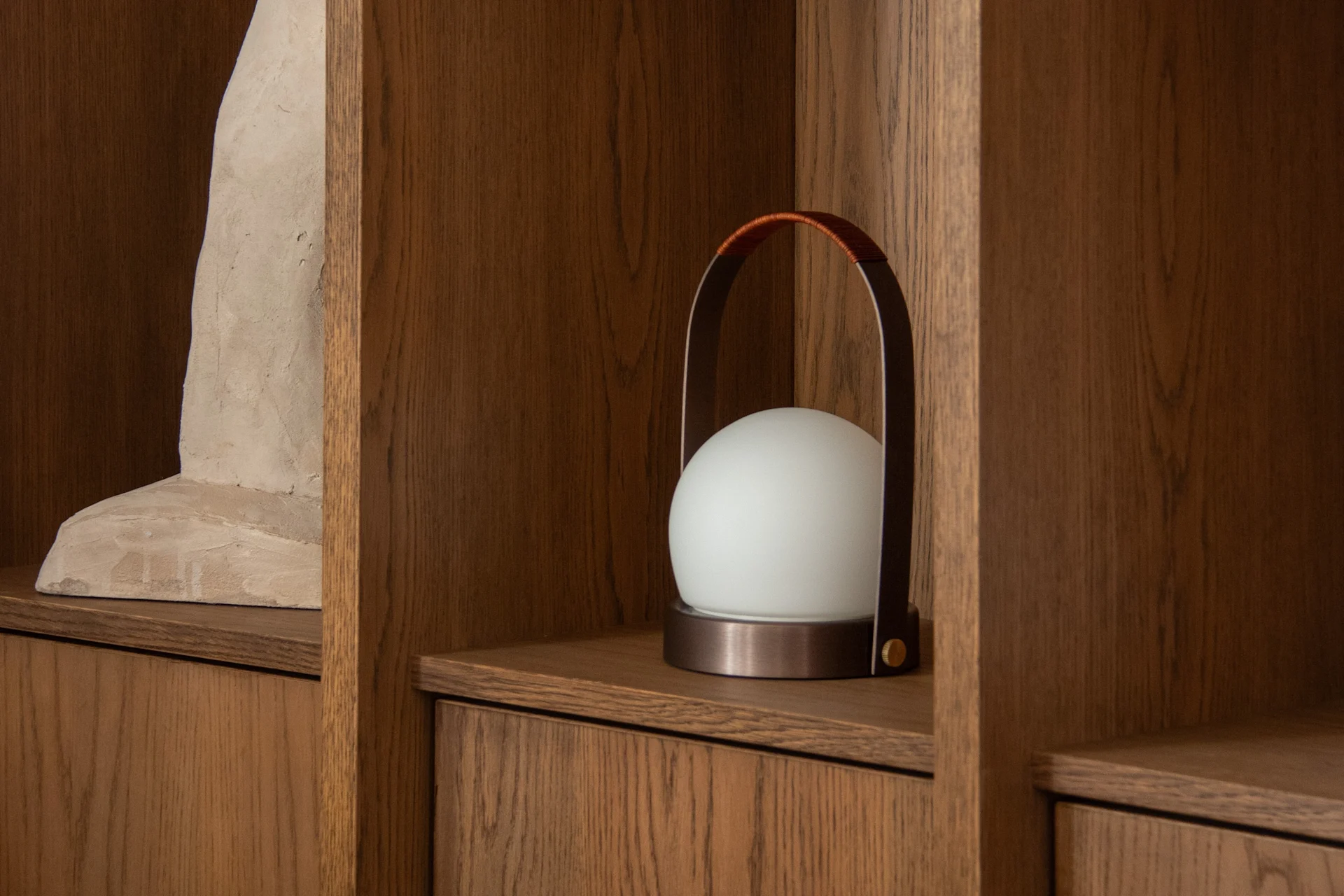 A modern white spherical lamp with a leather handle on a wooden shelf next to an abstract sculpture.