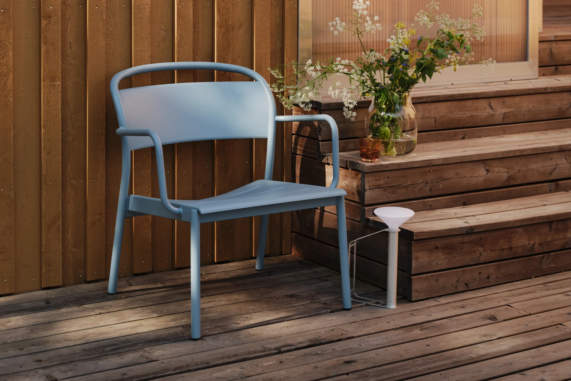 A light blue metal chair sits on a wooden deck next to a wooden wall, with a vase of wildflowers on a stair.