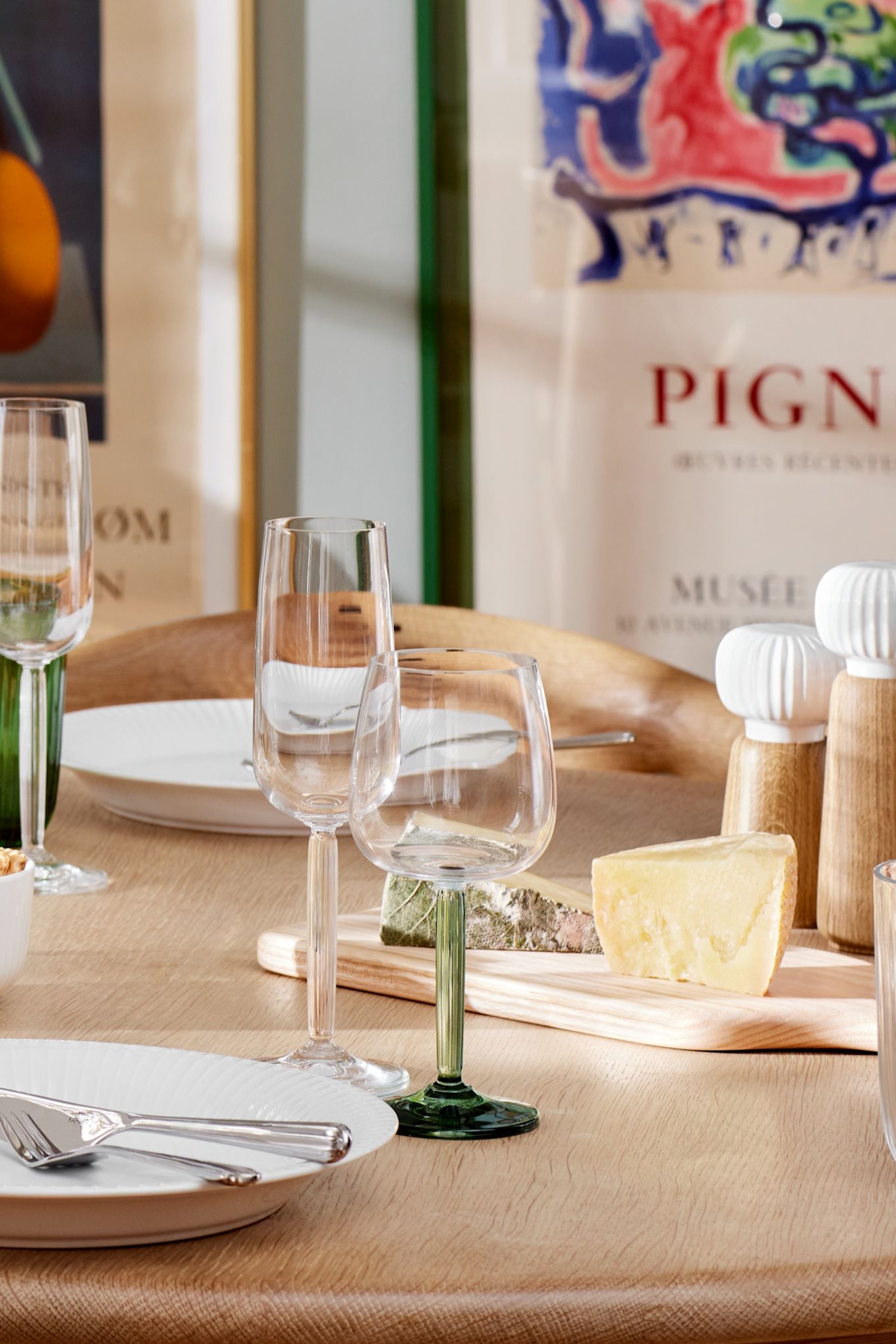 Hammshoi wine glasses with clear and green stems stand on a dining table in the sun.