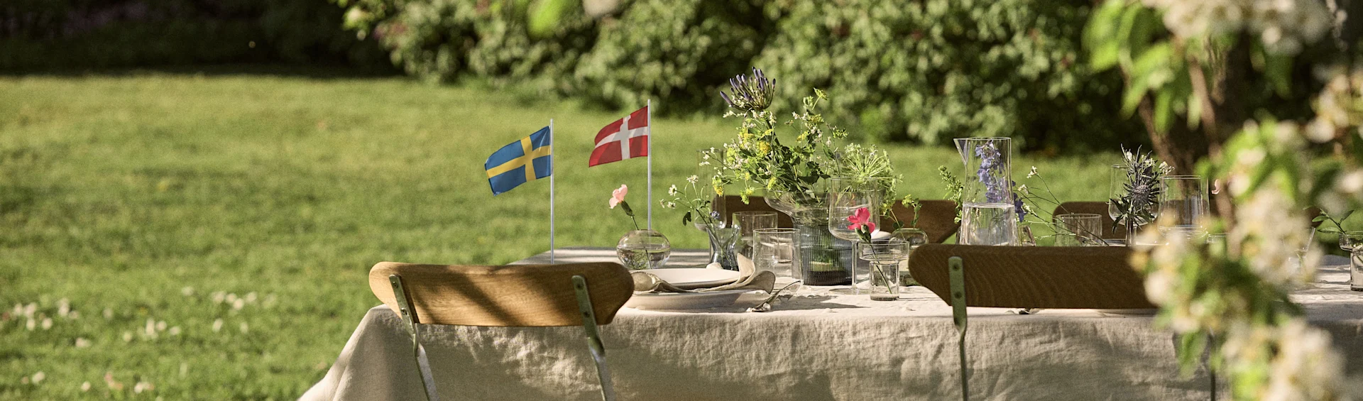 Outdoor table set with Swedish and Danish flags, wildflowers, and glassware in a sunny garden.