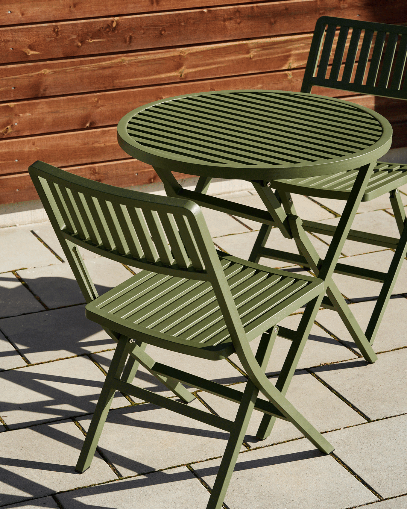 Green slatted outdoor table and two matching folding chairs on a patio with a wooden fence.