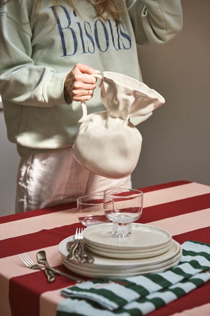 Person holding Crumple vase in white from Byon at a table set with a striped tablecloth in pink and burgundy. The Crumple vase has the shape of a crumpled paper bag, made of glazed ceramic.