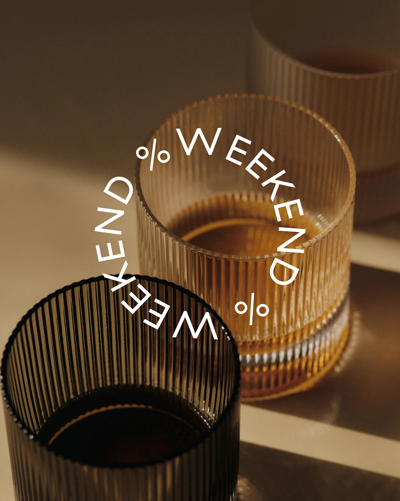 Three ribbed glasses, two filled with a dark liquid. A dark glass in the foreground, an amber glass behind it. Text: Weekend %.
