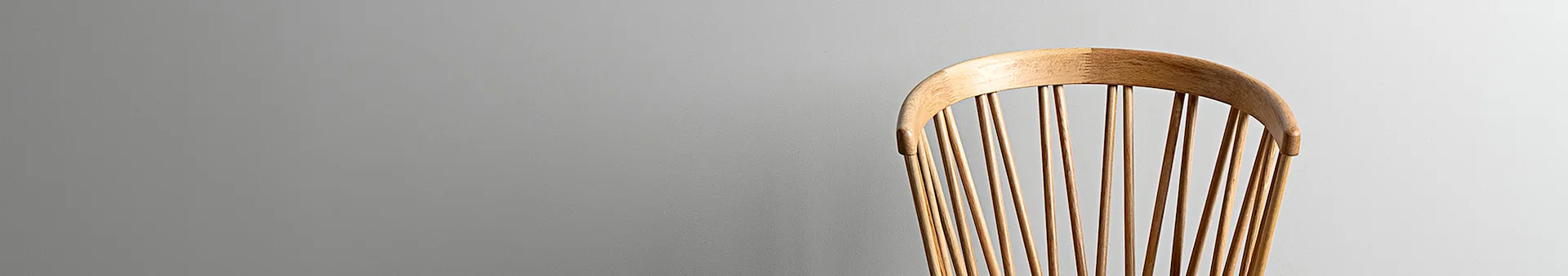 Back of a light wood spindle chair against a plain grey wall.