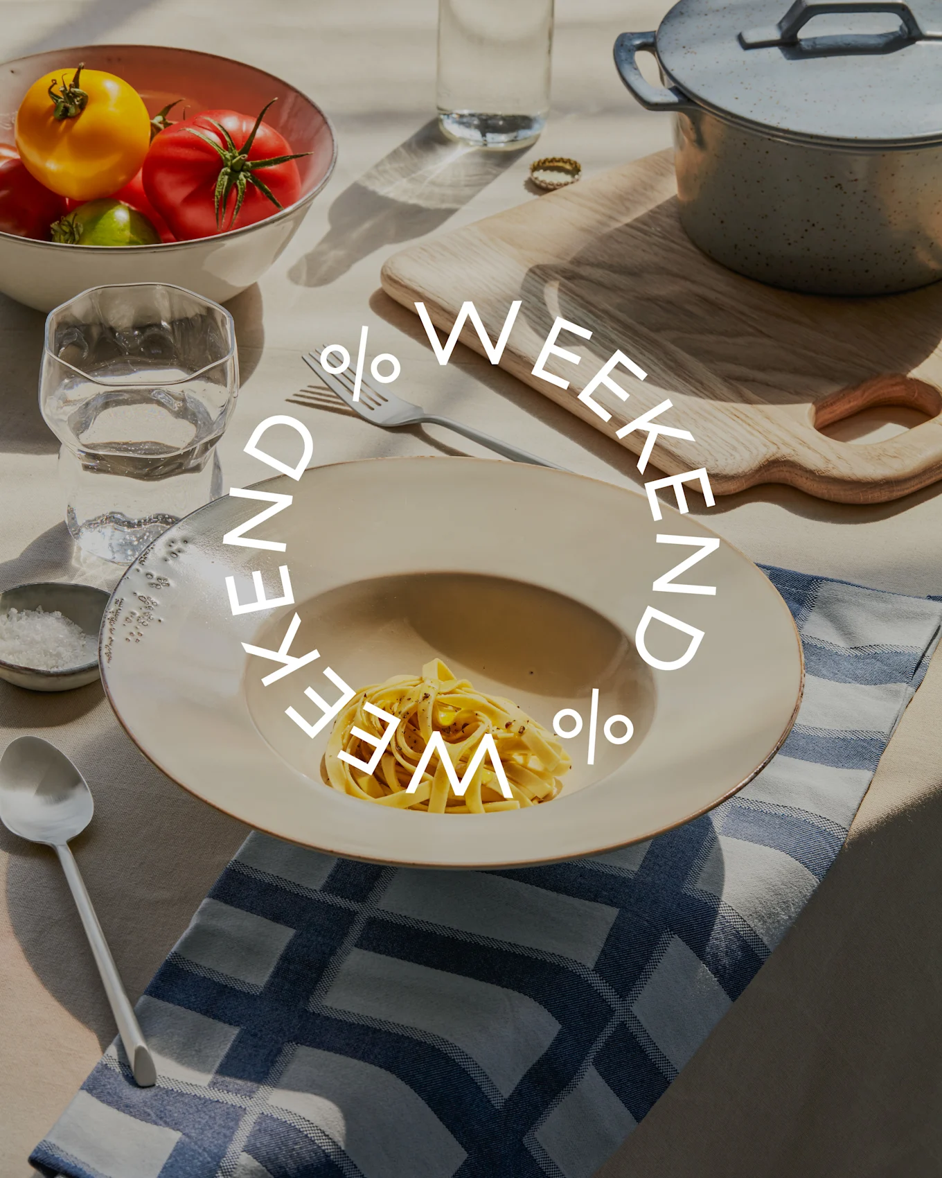 Table with pasta, a bowl of fresh tomatoes, water glass, and cooking pot. 'WEEKEND % WEEKEND %' text is overlaid.