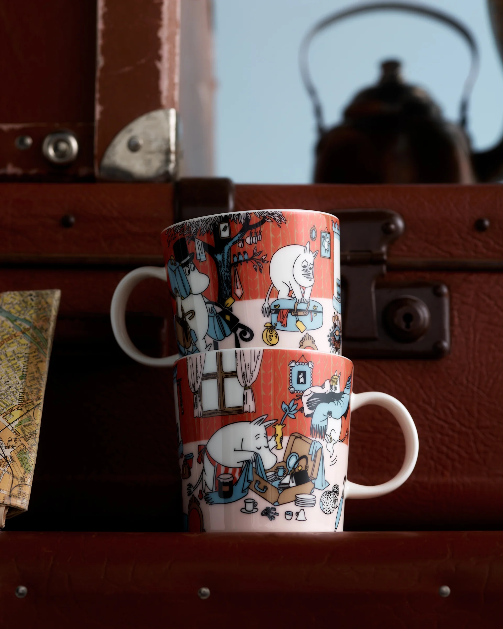 Two Moomin mugs with orange-red character illustrations, stacked in front of brown suitcases and a blurred map.