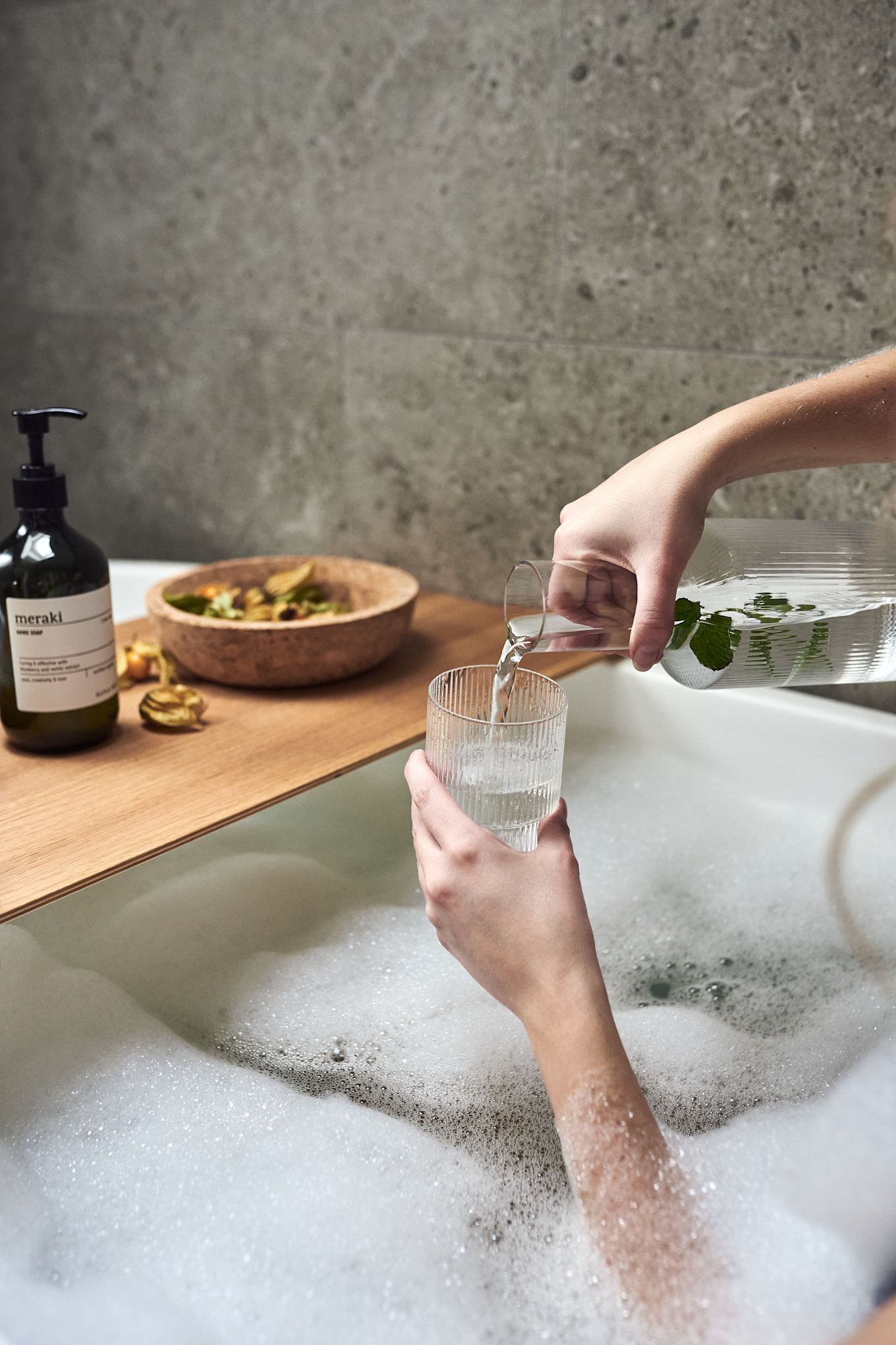 Hands pour water into a ripple glass from Ferm Living as part of a home spa night with a warm bubble bath. 