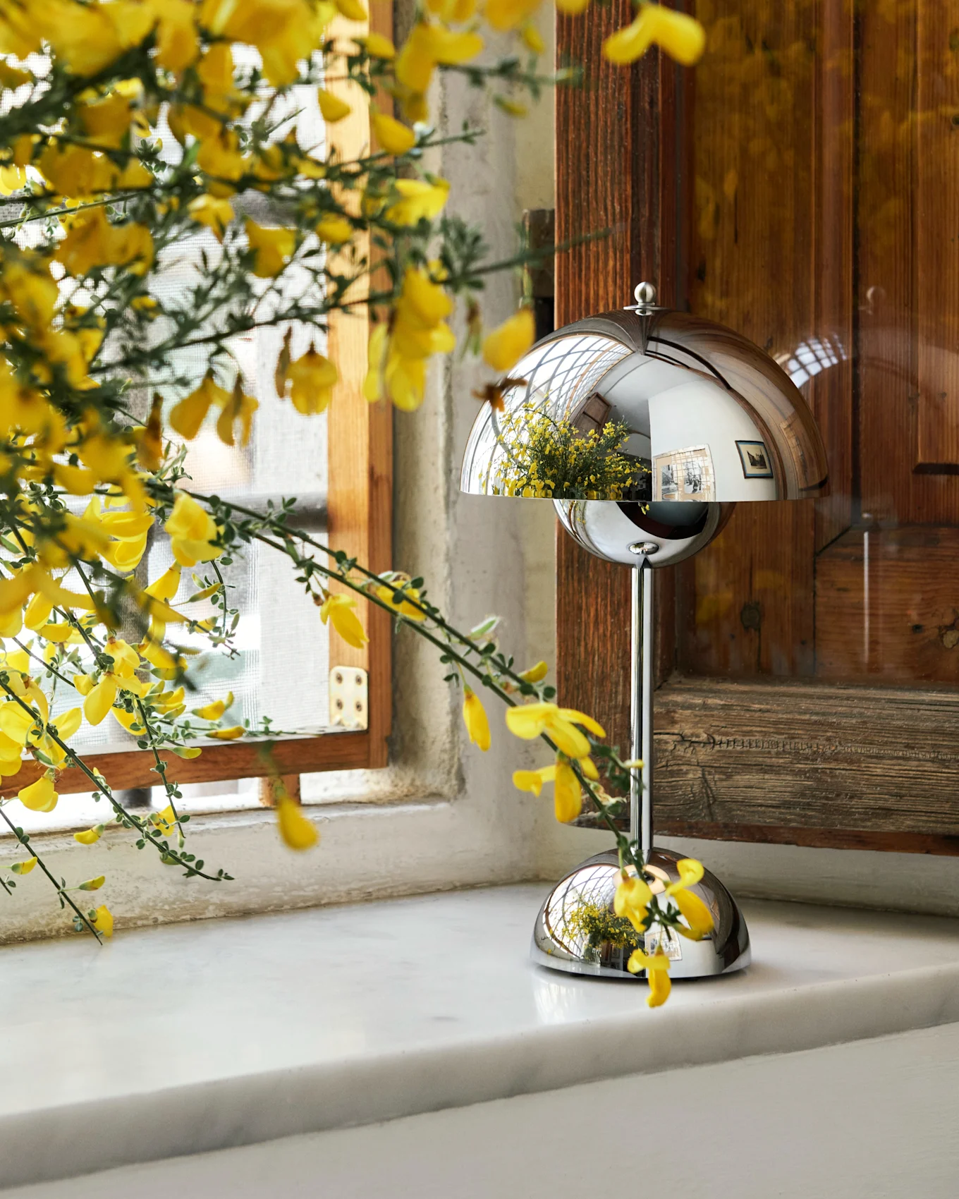 Yellow flowers in a vase on a marble windowsill next to a chrome mushroom-shaped lamp.