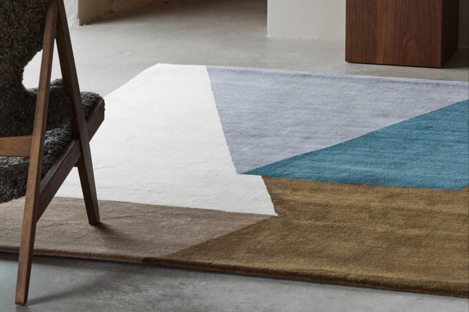 A geometric rug in white, light blue, turquoise, and brown on a concrete floor, with a wooden chair and a blue vase.