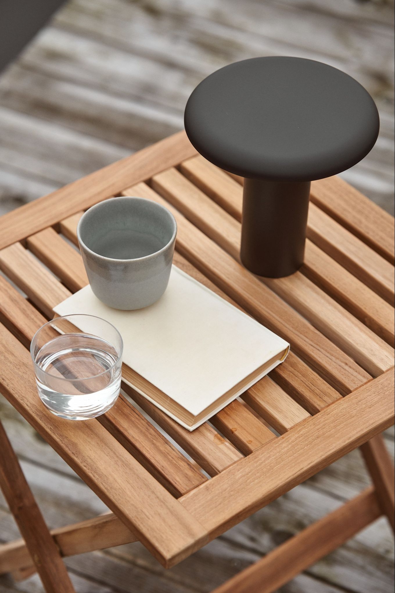 The Takku portable lamp from Artemide stands outside on a wooden table with a coffee cup and a glass of water. 