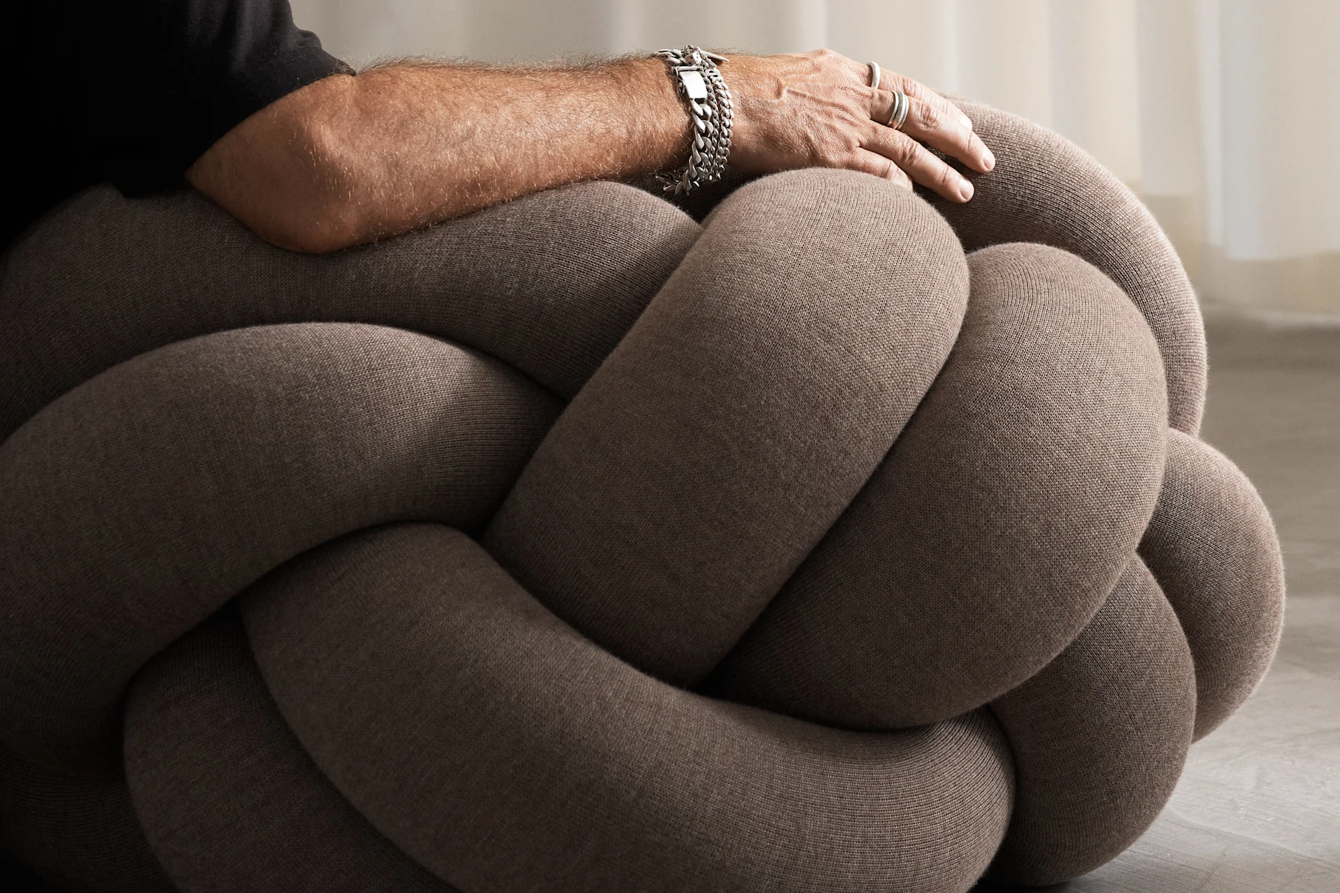 A man sits on top of the Knot Cushion XL in brown.