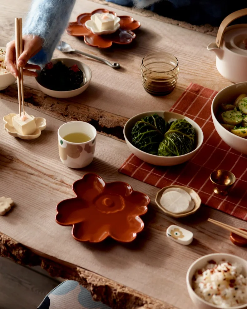 Woman with chopsticks serving food on a wooden table set with patterned ceramic dishes and a teapot.