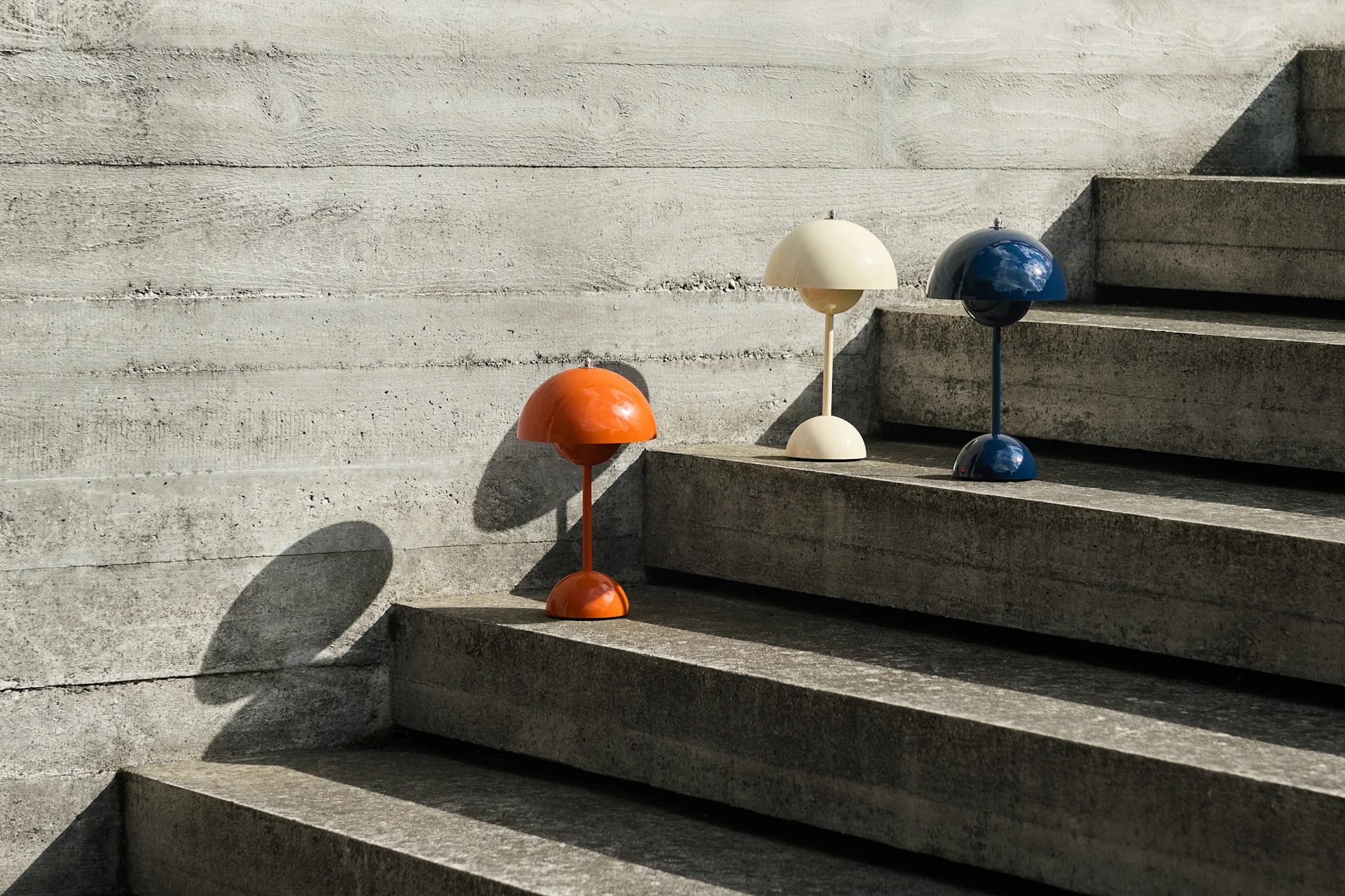 Three dome-shaped table lamps in orange, cream, and blue on concrete stairs next to a concrete wall, casting shadows.